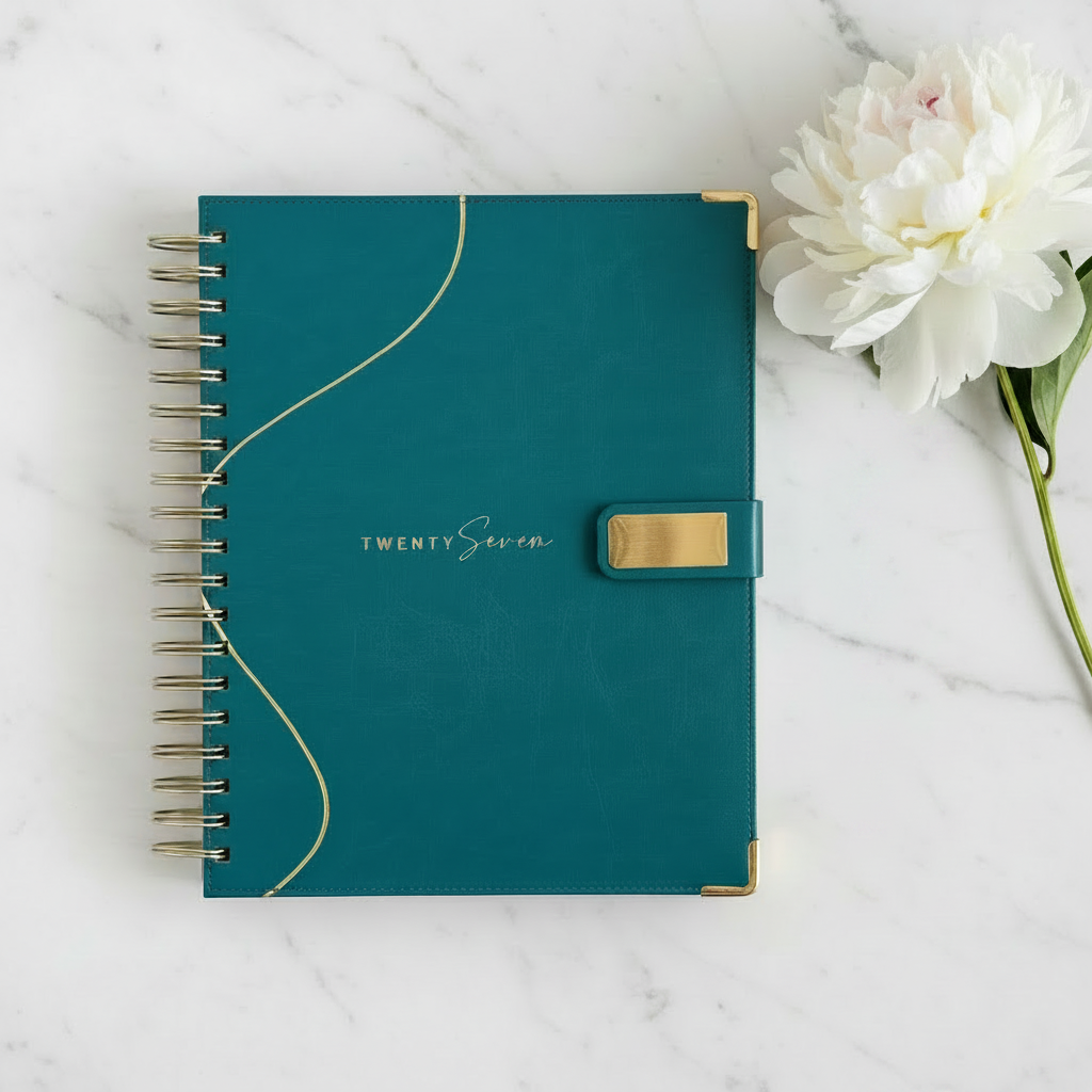 The 2027 teal planner with gold accents and a magnetic closure, on a white marble surface next to a white flower. The planner features gold twin wire coil binding on the left and the text 'TWENTYSeven' on the front cover.