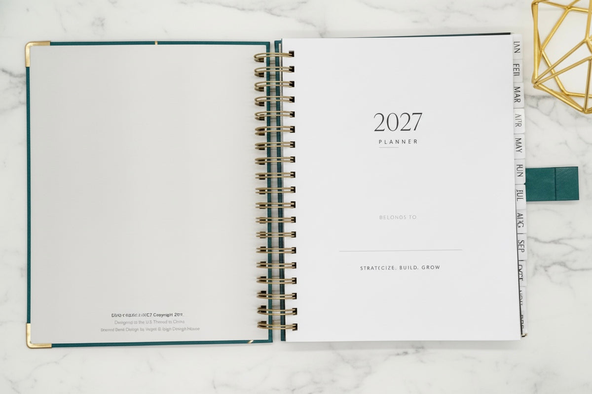2027 planner open layflat  on a marble surface with gold geometric decor. Planner is open to “this planner belongs to” page displaying twin wire coil, blinding metal reinforced corners and monthly tabs.