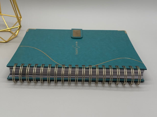 2027 Planner closed cover in teal with gold magnetic closure and coil binding, styled alongside a gold geometric decor piece.