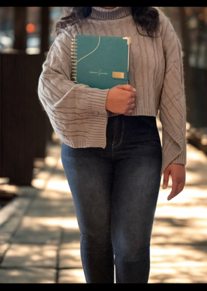 Person outdoors dressed in long sleeve grey sweater and dark pants holding 2027 teal planner with gold accents.