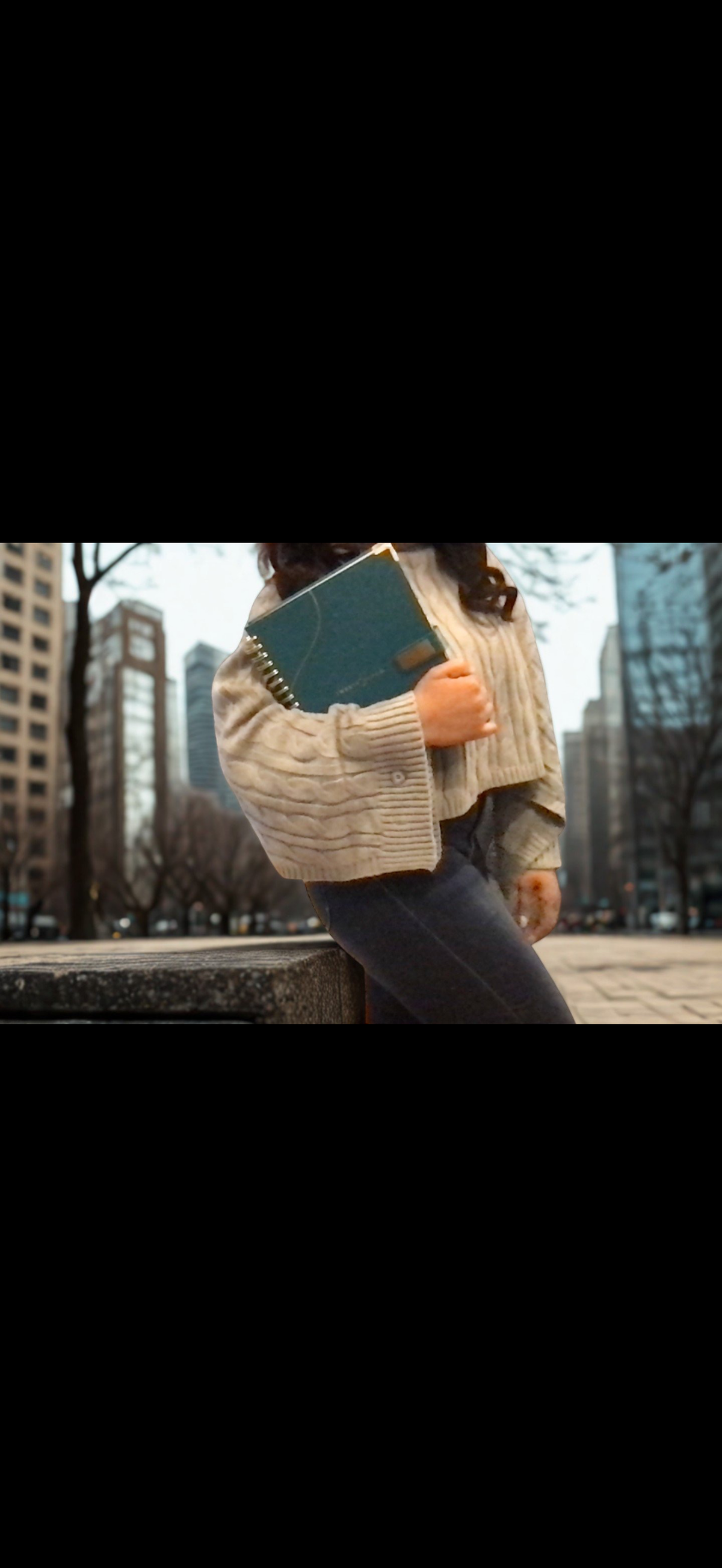 Person holding 2027 teal planner, wearing a long sleeve textured grey sweater in an urban setting with tall buildings in the background. Overlay text reads, Time. Steward it with intention.