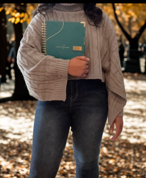 Person wearing a grey long sleeve sweater holding the teal 2027 planner with gold accents in an outdoor setting with trees and fallen leaves.