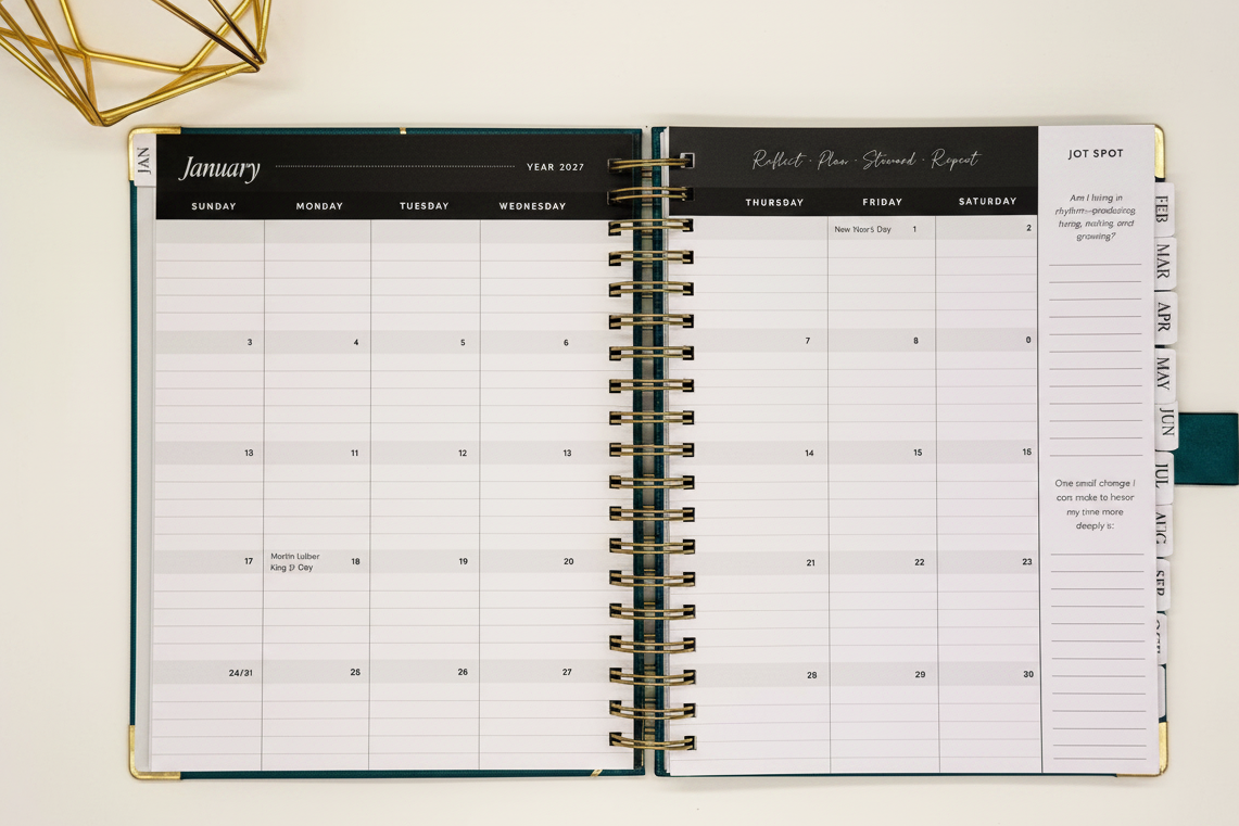 2027 Planner open to two-page monthly spread in spacious 7.5 x 10 format with tabbed months, coil binding, and styled alongside a gold geometric decor.