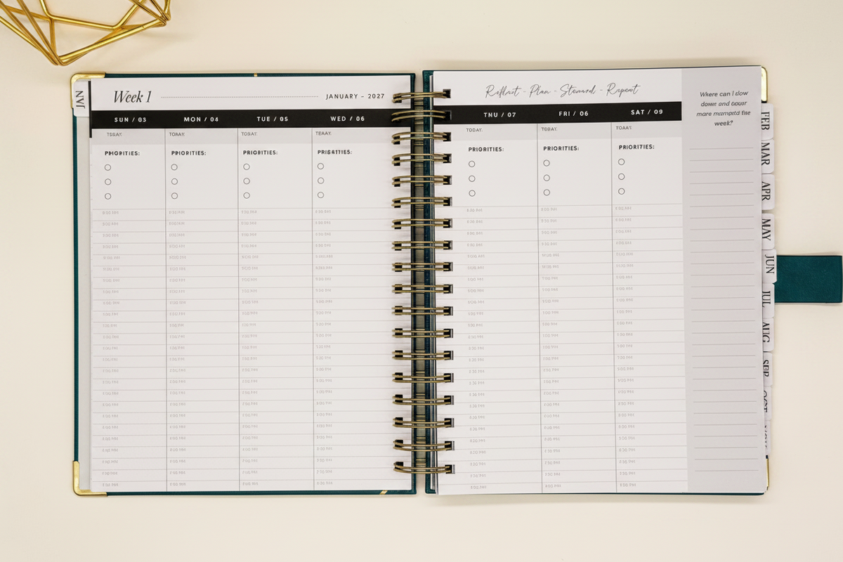 2027 Planner open to two-page weekly spread in spacious 7.5 x 10 format with tabbed months, coil binding, and styled alongside a gold geometric decor.