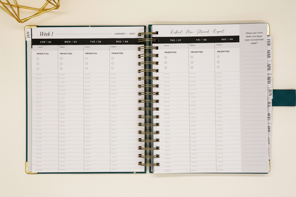 2027 Planner open to two-page weekly spread in spacious 7.5 x 10 format with tabbed months, coil binding, and styled alongside a gold geometric decor.