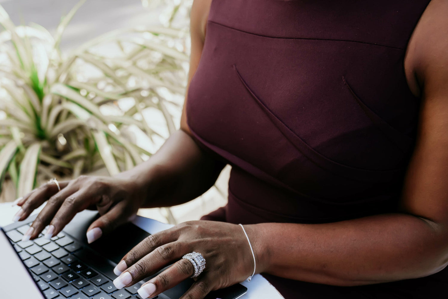 Person sitting, wearing a burgundy top  at white table, typing on laptop keyboard. Green plant in background.