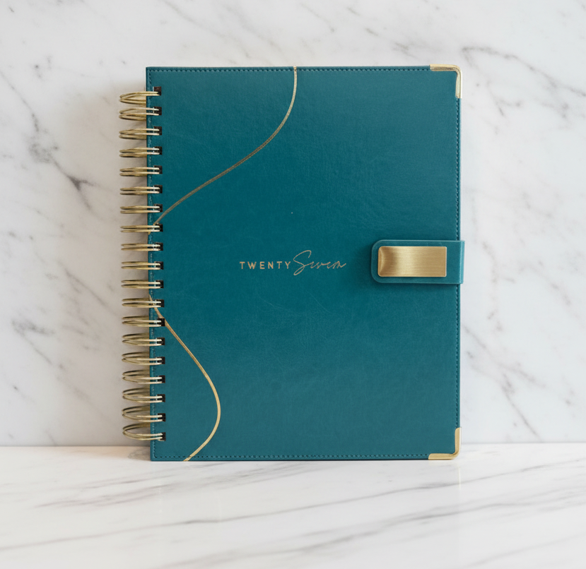 2027 teal planner, closed and upright with coil binding, metal reinforced corners and gold accent closure against white and grey marble background.