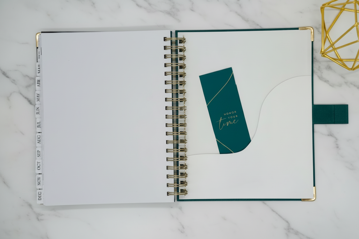 2027 Planner on Marble, open flat lay, displaying tabbed months, coil binding, pocket sleeve, and teal bookmark, styled alongside a gold accent.