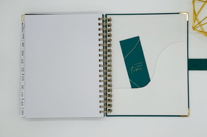 2027 Planner open flat lay showing tabbed months, back pocket sleeve with teal bookmark, reinforced gold corners, and coil binding, styled alongside a gold geometric decor. Cream background.